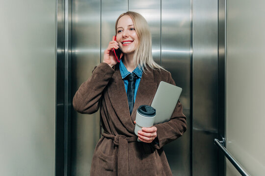 Businesswoman smiling while talking on phone with coffee and laptop in elevator