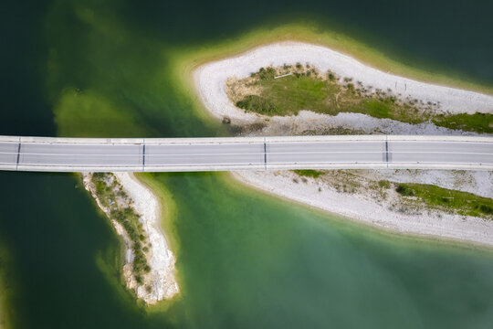 Aerial view of a bridge slicing through the emerald waters, connecting sandy shores touched by verdant patches, Sylvensteinspeicher, Bayern, Germany.