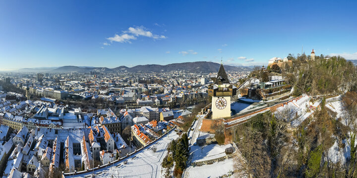 Aerial view of the snow-dusted cityscape with the iconic clock tower, contrasting with the hill topped by a historic fortress, Graz, Styria, Austria.