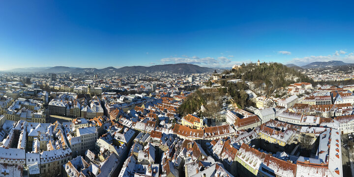Aerial view of snow-dusted rooftops and the iconic Schlossberg hill under a clear blue sky, framed by distant mountains, Graz, Styria, Austria.
