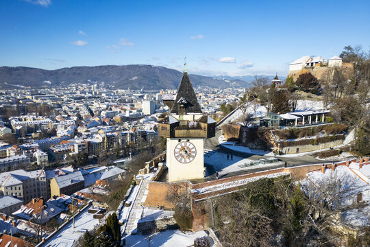 Aerial view of the iconic clock tower, Uhrturm, stands against the winter sky, amidst snow-covered rooftops and the distant mountains, Graz, Styria, Austria.