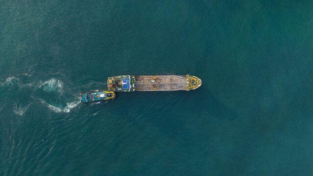 Aerial view of a tug boat nudging a ship across the blue-green waters, leaving white wakes like fleeting brushstrokes, Tambon Map Ta Phut, Chang Wat Rayong, Thailand.