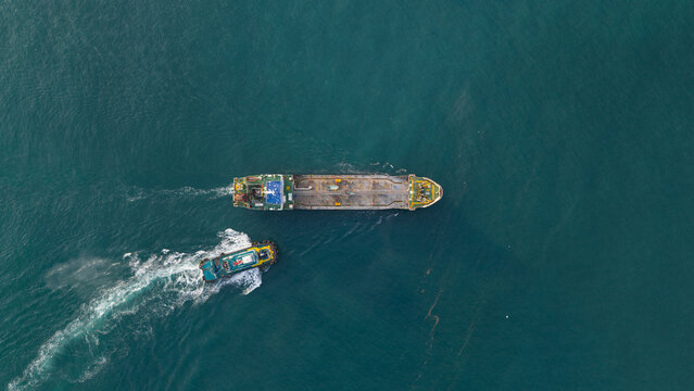 Aerial view of a large barge with a tugboat in tow, leaving a frothy wake across the tranquil, dark turquoise waters, Tambon Map Ta Phut, Thailand.