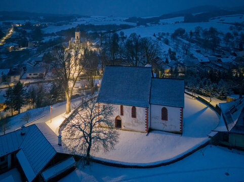 Aerial view of St Francis of Assisi Catholic Church under a blanket of snow, glowing softly against the fading light of dusk, Poniky, Banska Bystrica region, Slovakia.