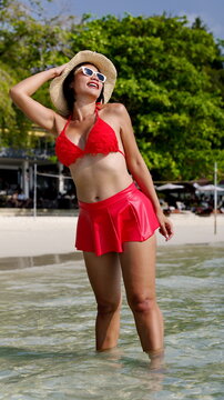 Asian woman in a red bikini top and matching mini skirt stands in the ocean while adjusting her straw hat and smiling