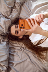 Young woman scrolling on the phone while lying in bed. Concept of social media content creation,...