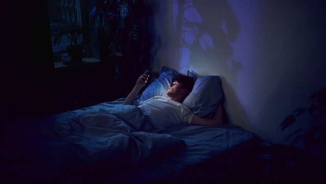 Young man lying in bed using smartphone late at night. Concept of streaming services, social media scrolling, ecommerce browsing, digital subscriptions and extended screen time.