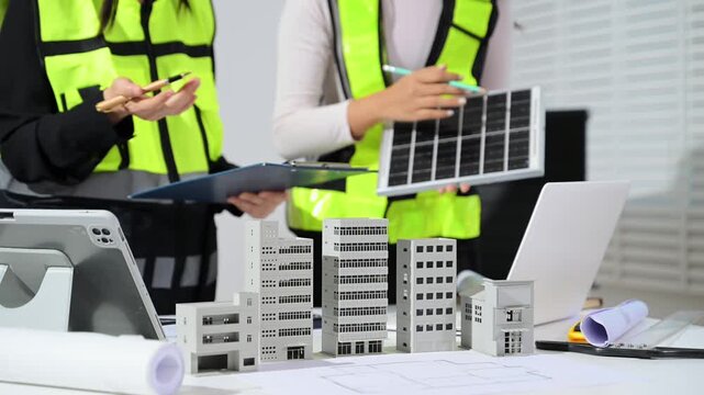 Construction engineers in safety vests reviewing blueprint and house model, using tablet and walkie talkie for project planning, site inspection