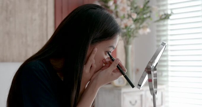 asian young woman drawing eyeliner in front of mirror with concentration during personal beauty routine in bright bedroom showing quiet selfcare moment and indoor lifestyle with soft morning light