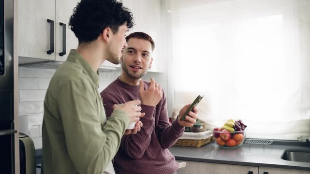 Lovely young gay couple enjoying a morning conversation in the kitchen, one man drinking coffee while the other shows him something on his phone