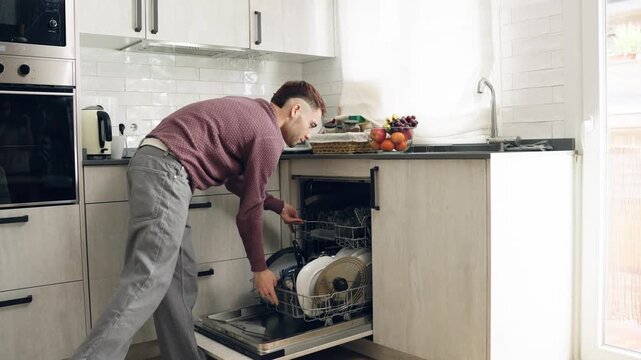 Young man doing household chores, taking clean dishes out of the dishwasher. Housekeeping and daily routine in a modern bright kitchen