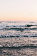 Calm ocean waves with white foam roll towards the shore under a soft pink and peach twilight sky. The minimalist seascape captures a tranquil and meditative atmosphere during the golden hour.