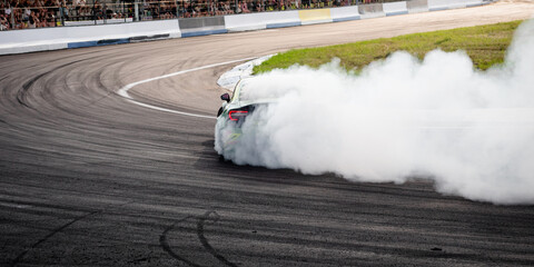 Green drift car entering a turn with smoke. Dynamic rear-angle shot of a modified red sportscar as it slides through a track corner. Unfocused grandstands in the background. © andrbk