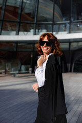Obraz premium Smiling businesswoman in sunglasses standing near corporate building outdoors