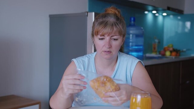 A woman sitting at a table in front of a kitchen background takes a confectionery product out of its packaging and tastes