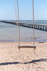 Fototapeta premium Wooden swing hanging from ropes on sandy beach with a pier extending into the calm sea under a clear blue sky