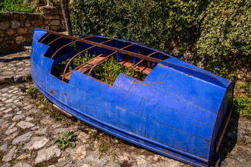 An old, rusted blue fishing boat hull rests upside down on a cobbled ground patch, partially overgrown with green ivy and small plants, bathed in bright sunlight in Ohrid, North Macedonia.