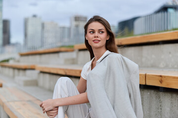 Young woman sitting outdoors in casual white outfit with light jacket, modern urban background,...