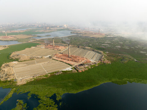 Aerial view of brick kilns casting long shadows across the landscape, contrasting with the lush green vegetation along the water's edge, Aminbazar, Dhaka Division, Bangladesh.