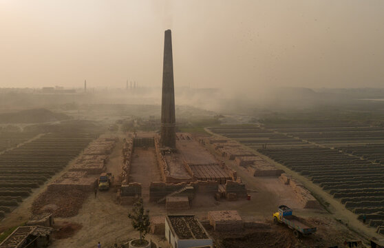 Aerial view of a towering brick chimney dominates the skyline amidst earth-toned brick fields under a hazy sky, Aminbazar, Dhaka Division, Bangladesh.