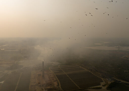 Aerial view of a hazy landscape where industrial chimneys belch smoke into the sky, and birds are flying over the land, Aminbazar, Dhaka Division, Bangladesh.