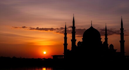 Obraz premium A breathtaking sunset paints the sky with fiery hues behind the majestic silhouette of a grand mosque, creating a serene and spiritual landscape at dusk as reflections dance on the water