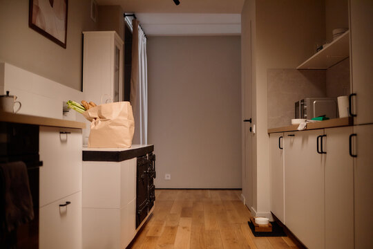 Modern kitchen interior showing paper grocery bag with vegetables on countertop, wooden floor, cabinets, microwave and kitchen appliances, empty hallway in background