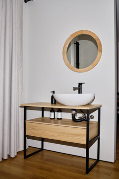 Modern bathroom vanity featuring round wooden mirror, vessel sink, exposed plumbing, liquid soap dispenser and lotion bottle standing on wooden countertop against wall