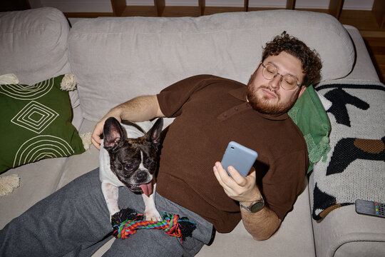 Caucasian young adult man reclining on sofa holding smartphone in right hand while French Bulldog sitting on lap yawning, colorful rope toy resting near dog, television remote visible