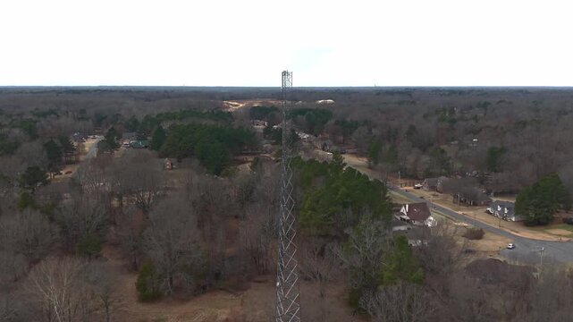 Cinematic drone footage of a tall steel radio tower rising above a rural landscape with surrounding trees, fields, and a nearby country home.