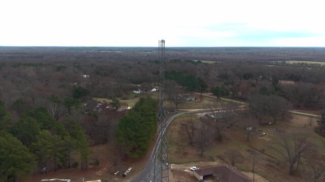 Cinematic drone footage of a tall steel radio tower rising above a rural landscape with surrounding trees, fields, and a nearby country home.