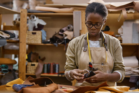 Black young adult woman working with leather pieces in workshop, using hands to inspect material, wearing glasses and apron, surrounded by tools and leather supplies on workbench