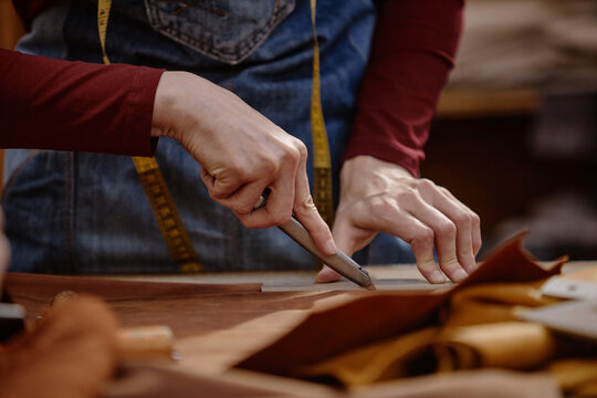 Caucasian woman cutting leather with utility knife on workbench in workshop, hands visible holding tool and guiding material, measuring tape hanging around neck