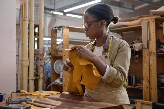 Black young adult woman working in leather workshop holding large piece of leather inspecting material wearing apron standing at workbench surrounded by leather crafting tools and supplies