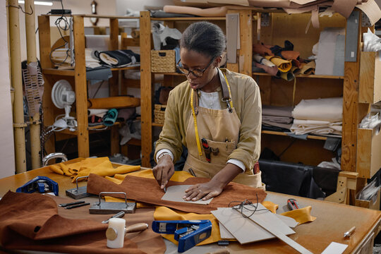 Black woman young adult working in leather workshop cutting brown leather fabric on worktable using ruler and utility knife surrounded by crafting tools and materials in organized workspace
