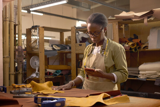 Black woman young adult standing in leather workshop using smartphone while working with leather pieces on workbench, surrounded by tools and materials, focusing on craft process