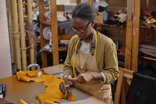 Black woman young adult working in leather workshop using hammer and crafting leather piece on workbench, wearing apron and measuring tape around neck, focused on precise handwork