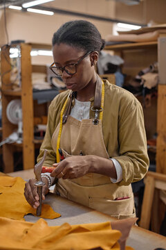 Black young adult woman working in leather workshop using hand tool to cut leather hide, wearing apron and glasses, focused on precise craftsmanship at workbench, measuring with tape