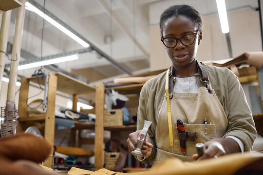 Black woman young adult working in leather workshop using tools to craft leather goods, standing at workbench with shelves and materials in background, focused on precise hand movements