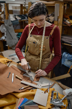 Young adult Caucasian woman working in leather workshop, using hand tool to cut leather on workbench, surrounded by crafting tools and materials, focused on precise craftsmanship