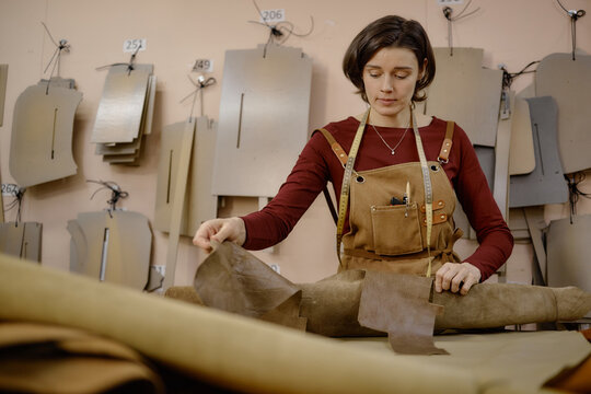 Caucasian young adult woman working in leather workshop handling brown leather material on worktable, measuring patterns hanging on wall in background, focused on crafting process