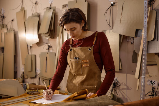 Young adult Caucasian woman working in leather workshop, standing at workbench writing notes on paper, surrounded by leather patterns and tools, focused on craftsmanship process