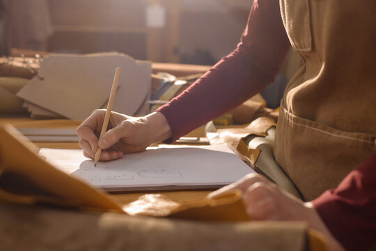 Caucasian woman sketching leather pattern on paper at workshop table, using pencil with leather pieces and crafting materials surrounding workspace, focusing on design process