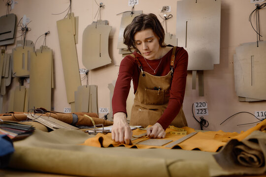 Caucasian young adult woman working in leather workshop cutting fabric with ruler and measuring tape, surrounded by leather pieces and pattern templates hanging on wall in background