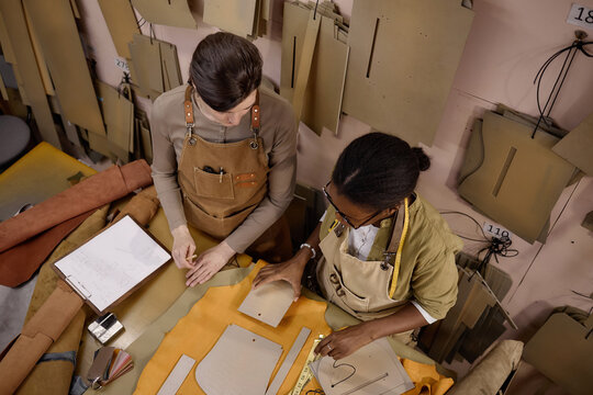Caucasian young adult woman and Black young adult woman working together in leather workshop, cutting and measuring leather pieces on worktable, using templates and sketches