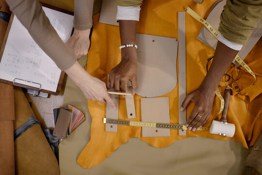Caucasian woman and Black woman collaborating on leather crafting project, measuring and arranging leather pieces on workshop table, using ruler, tape measure, and pattern templates