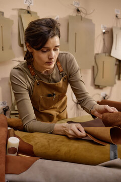 Caucasian woman working in leather workshop inspecting brown leather hide on worktable surrounded by pattern templates and crafting materials, focused on quality control process