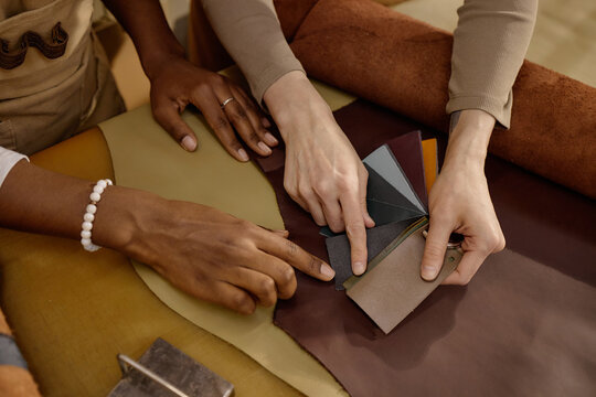 Caucasian woman and Black woman selecting leather swatches on worktable, hands arranging material samples in workshop, collaborating on design choices for handcrafted leather goods
