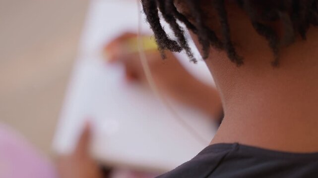 black teen sketching on tablet wearing earbuds and seated on pink chair cropped backofhead view shows textured locks, nape and shoulder while fingers move across screen, intimate creative study