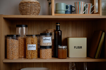 Fototapeta premium Kitchen shelf displaying transparent containers filled with pasta, oatmeal, lentils, glass bottle of oil, jar of honey, metal flour tin, stacked mugs and woven basket on upper shelf